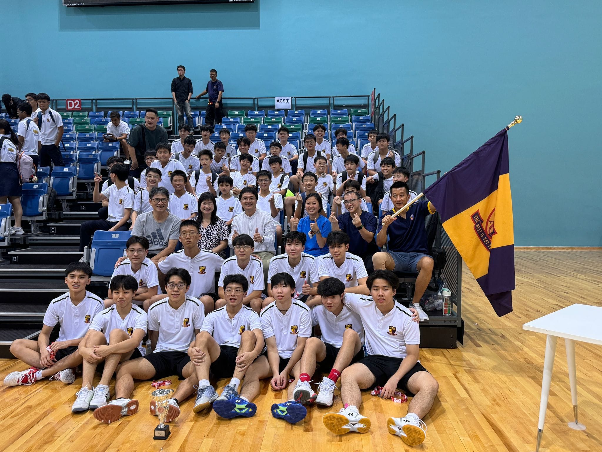 A large group of students in white uniforms poses in a sports hall with tiered spectator seating, with one student holding a school flag in the center of the group.